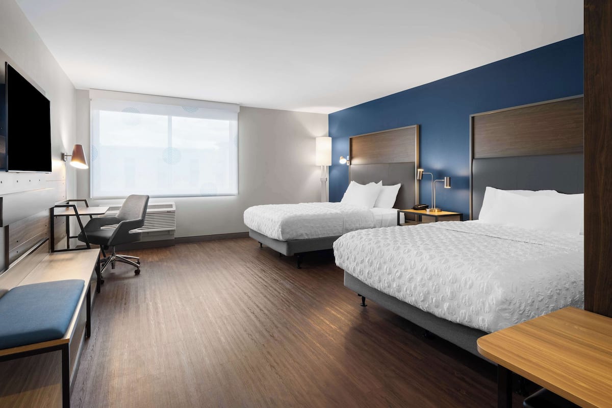 A hotel room with two beds, a desk, a chair, a TV, and a window.