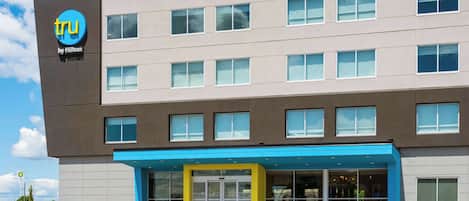 A modern hotel building with a prominent 'tru by Hilton' sign, multiple windows, and a blue entrance area.