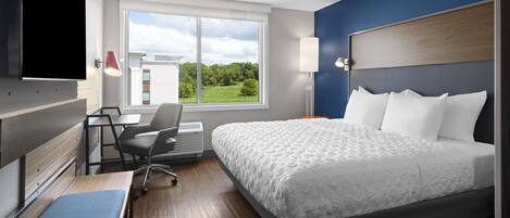 A hotel room with a large bed, a desk with a chair, a TV mounted on the wall, and a window with a view of greenery.