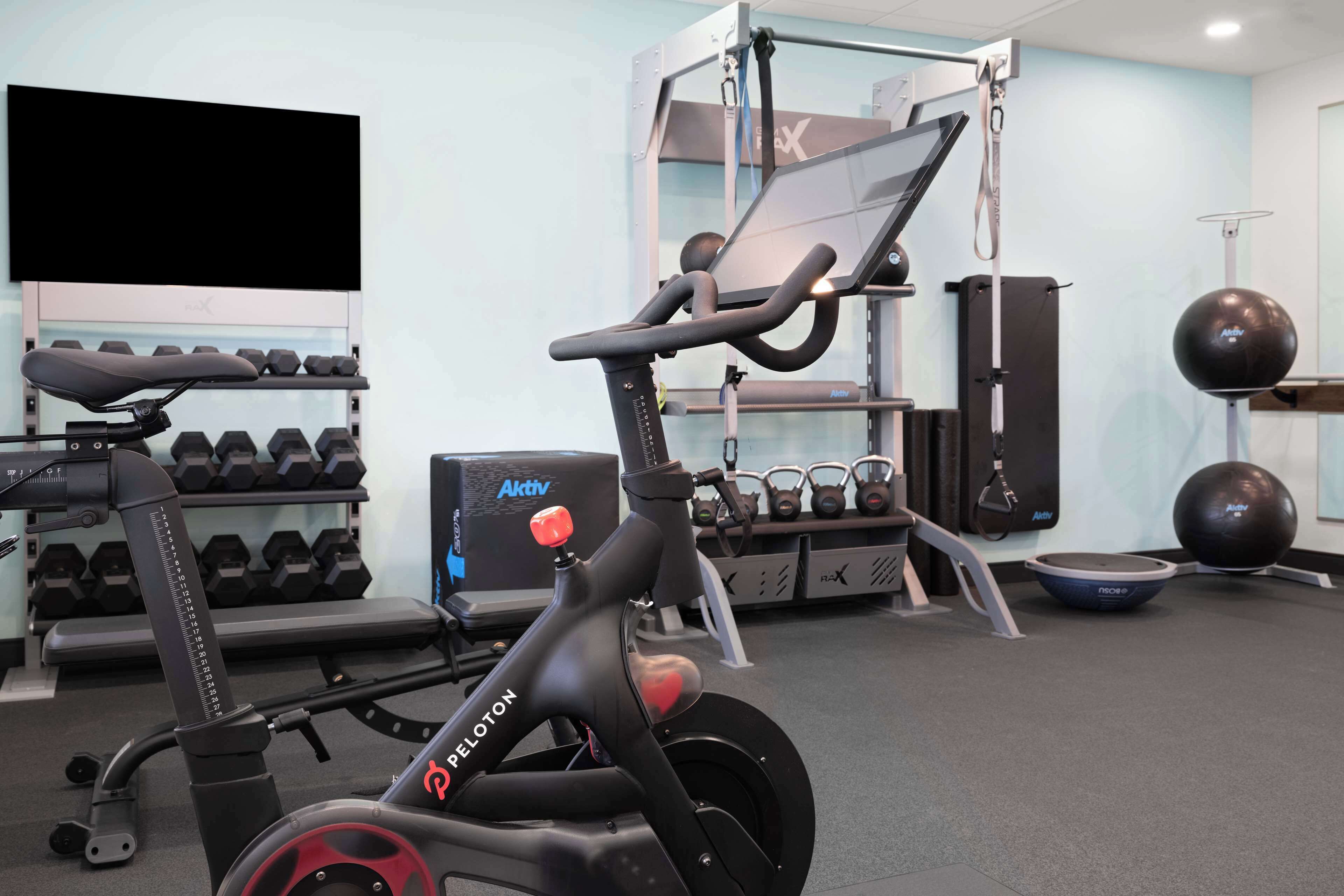 Una bicicleta estática Peloton en un gimnasio con mancuernas, kettlebells y pelotas de ejercicio.