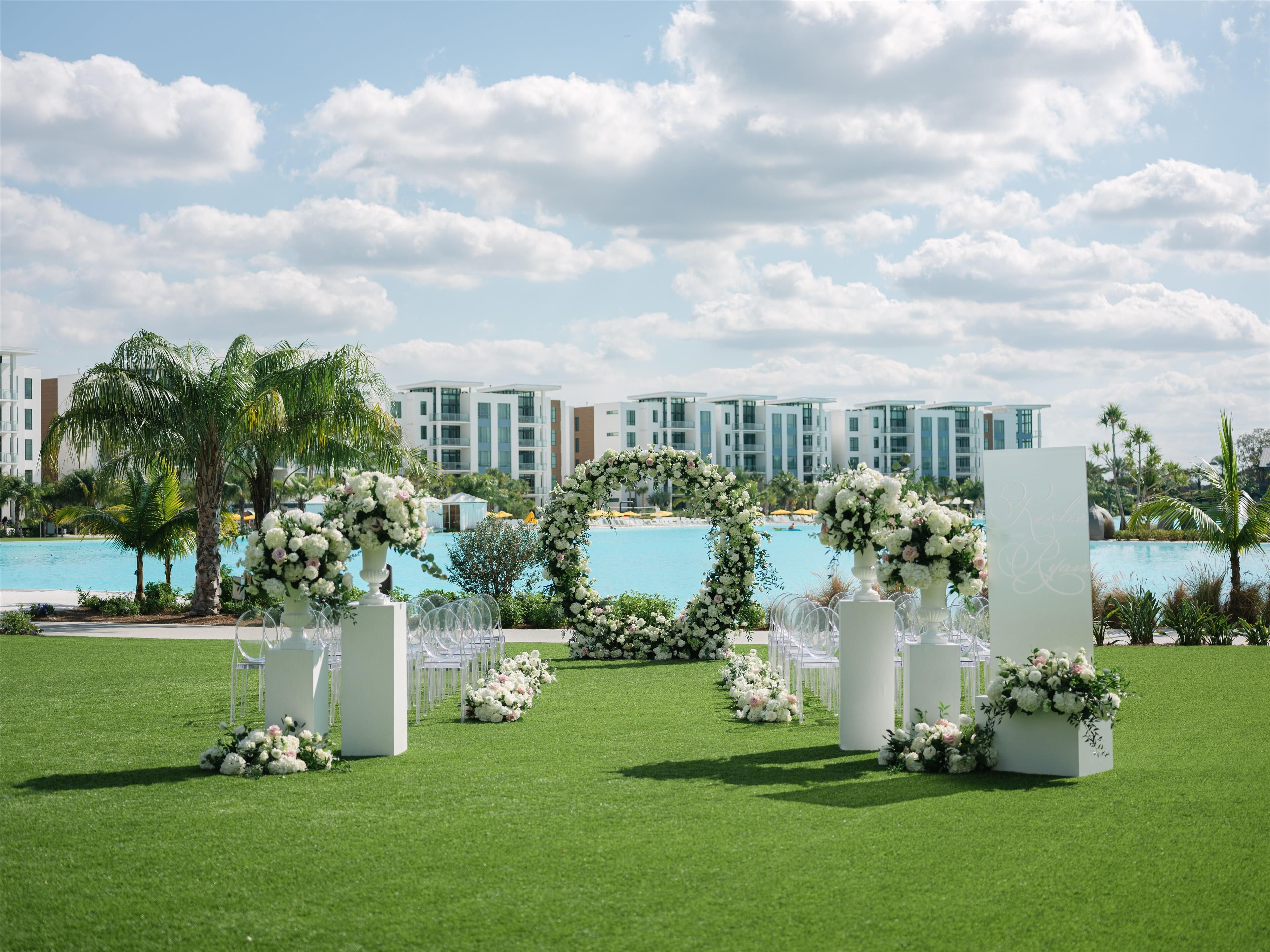 Ein Hochzeitszeremoniell mit weißen Blumarrangements und Stühlen, ein Pool und moderne Gebäude im Hintergrund.