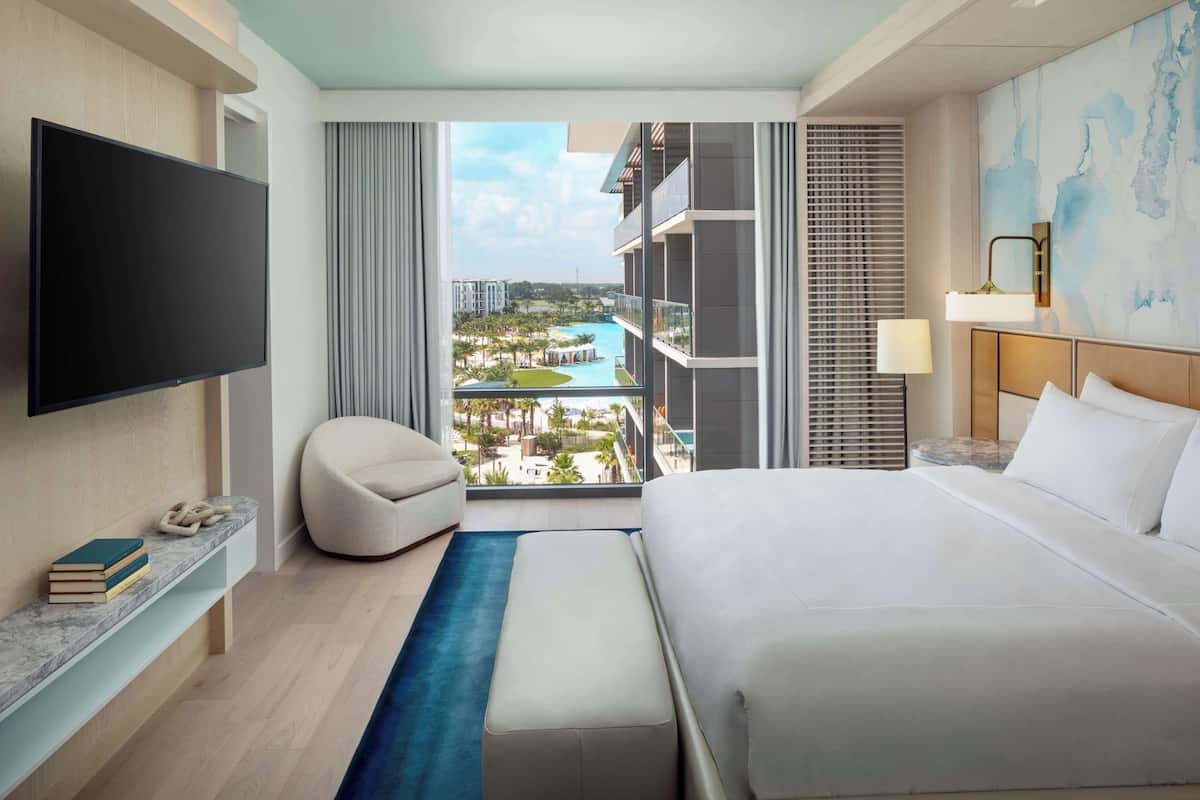 Une chambre d’hôtel moderne dotée d’un grand lit, d’un téléviseur à écran plat, d’une vue sur une piscine et des bâtiments, et d’un balcon avec une balustrade.
