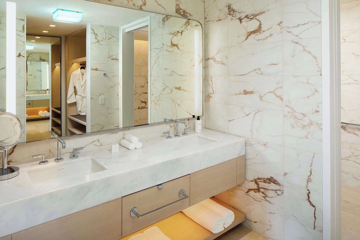 A bathroom with a marble wall, a large mirror, a double sink, and a towel rack.