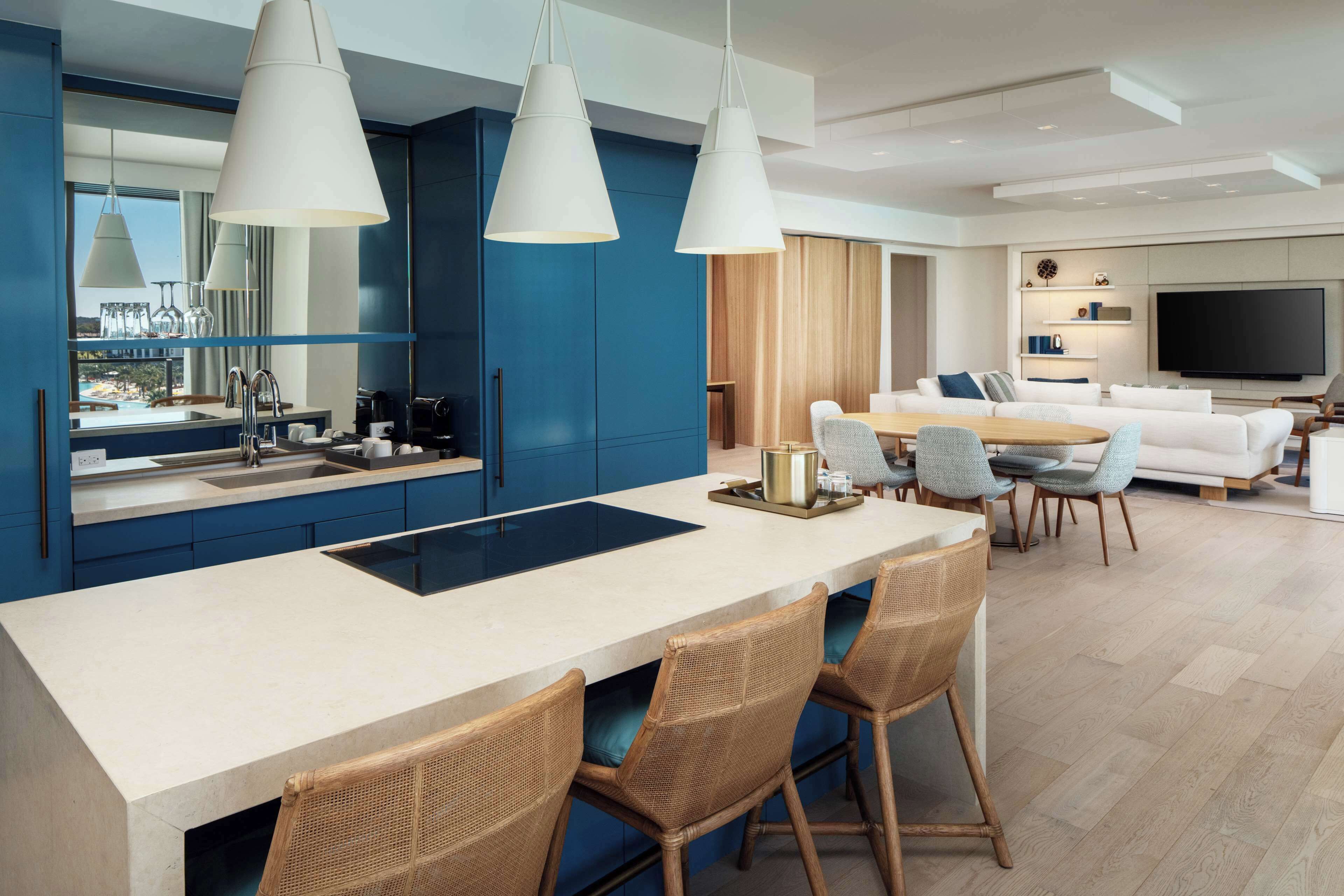 A modern kitchen with a central island, dining area with wicker chairs, a large flat-screen TV, and a sofa in the living room.