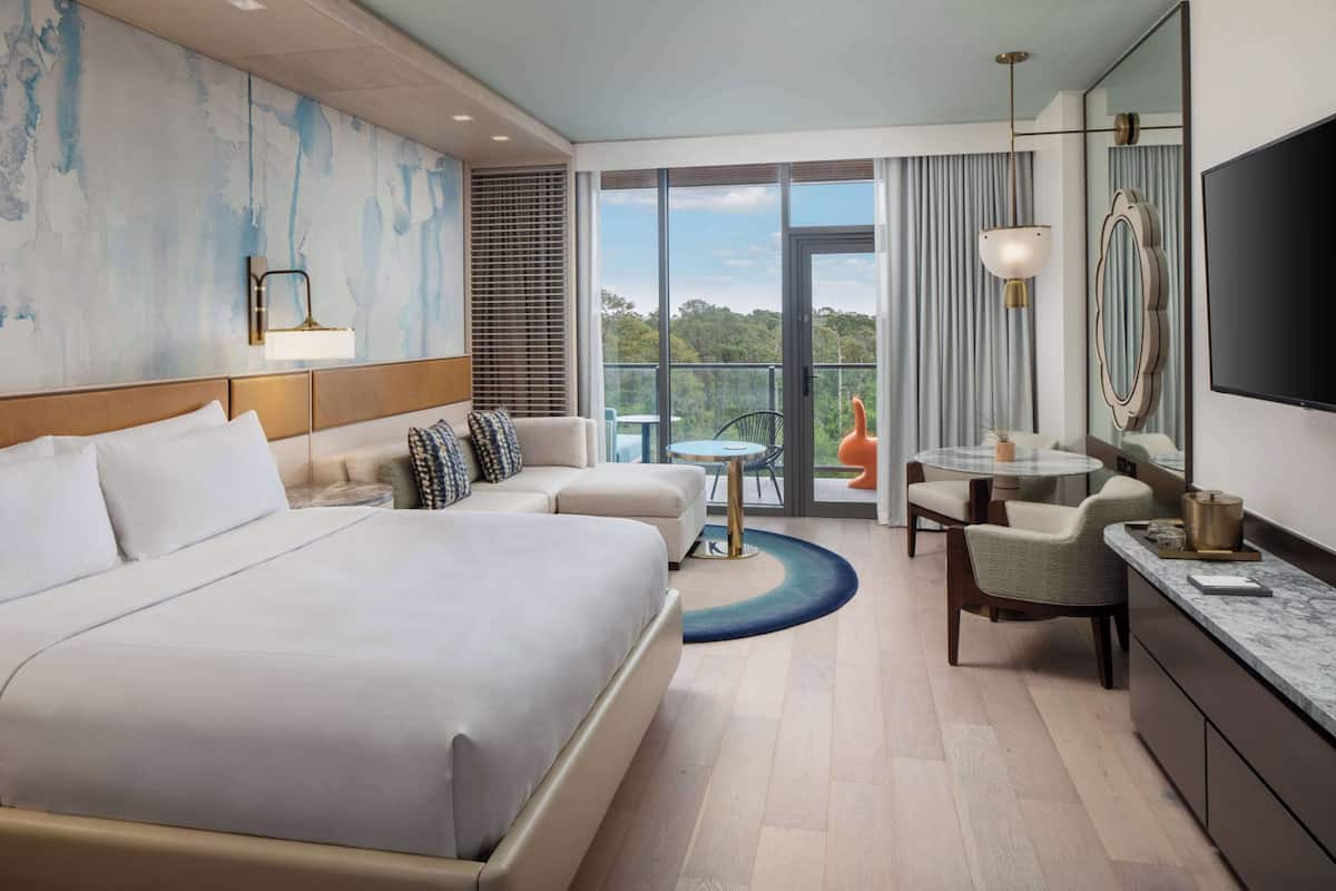 Une chambre d’hôtel avec un grand lit, un coin salon avec des fauteuils, une petite table, une télévision et un balcon donnant sur les arbres.