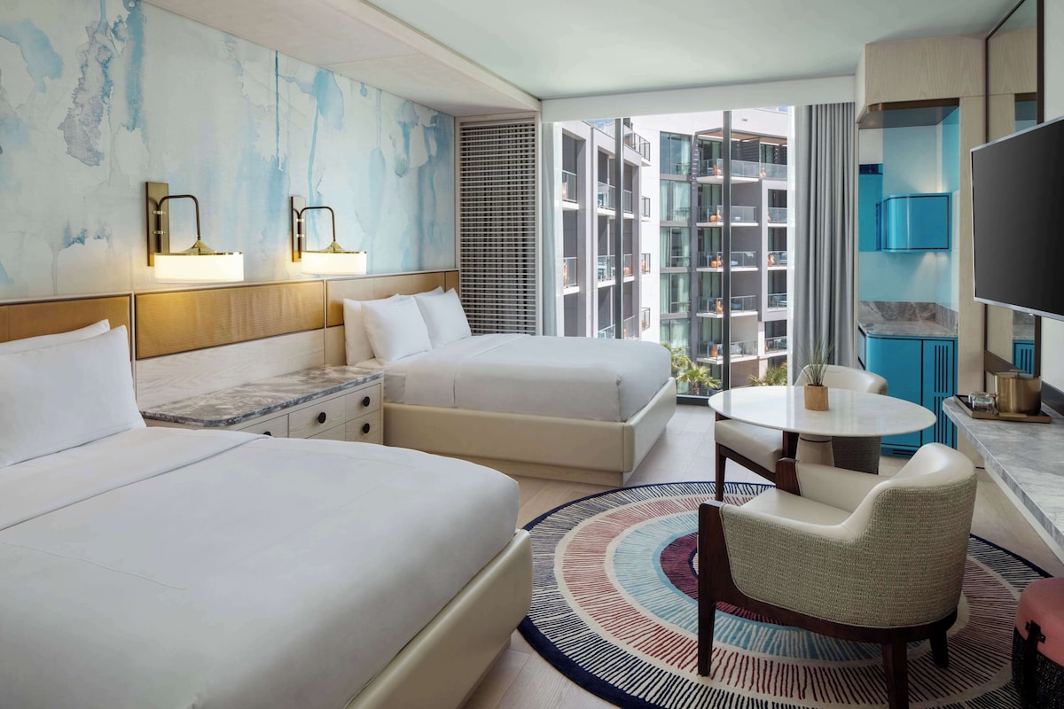 A hotel room with two beds, a dining table, a TV, and a large window with a view of high-rise buildings.