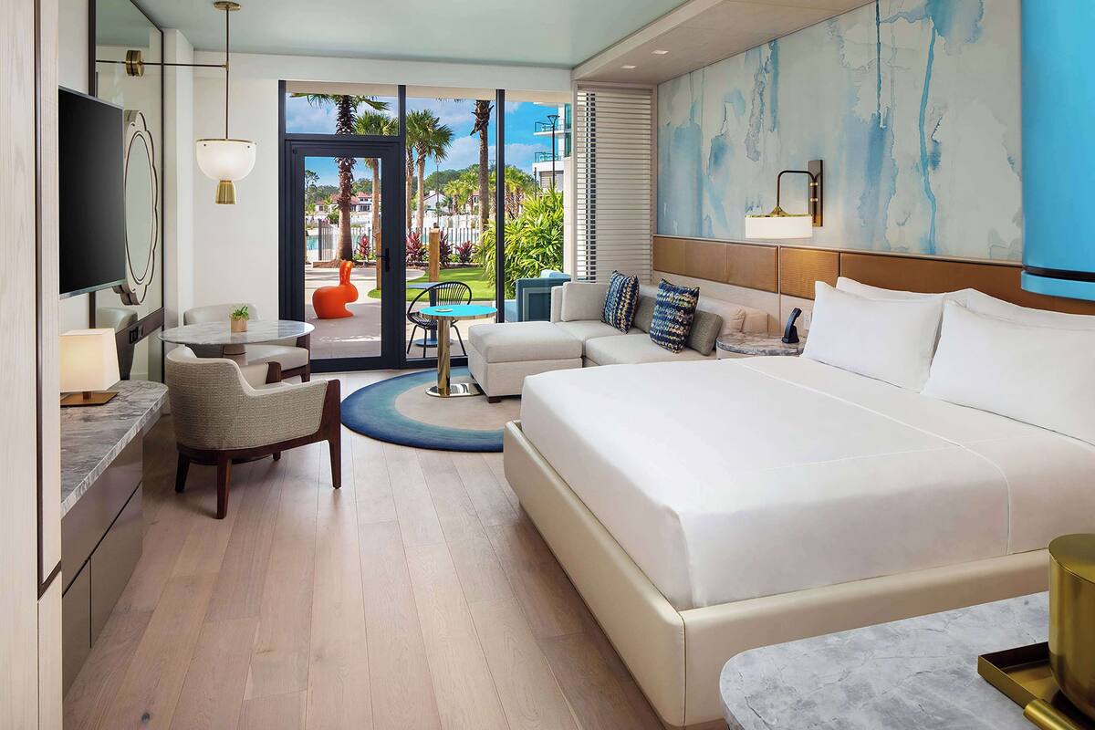 Une chambre d’hôtel moderne dotée d’un grand lit, d’un canapé, d’un coin repas avec une table et des chaises, et offrant une vue sur les palmiers et les bâtiments environnants.