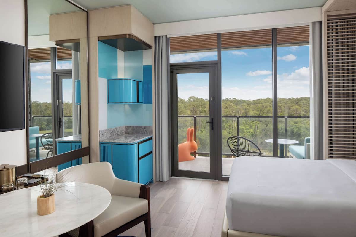 Une chambre d’hôtel moderne dotée d’un grand lit, d’un coin repas avec une table et des chaises, d’un coin cuisine avec des meubles bleus et d’un balcon offrant une vue sur les arbres.