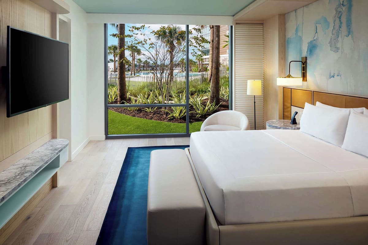 Une chambre d’hôtel moderne dotée d’un grand lit, d’un téléviseur à écran plat, d’un banc, d’un bureau avec une lampe et offrant une vue sur une piscine et un espace verdoyant.