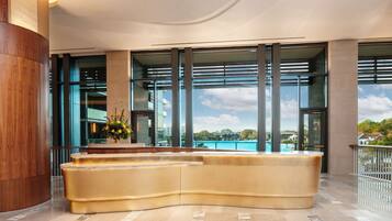 A modern reception area with a curved reception desk, large windows offering a view of the pool and surrounding landscape, and a wooden column on the left.