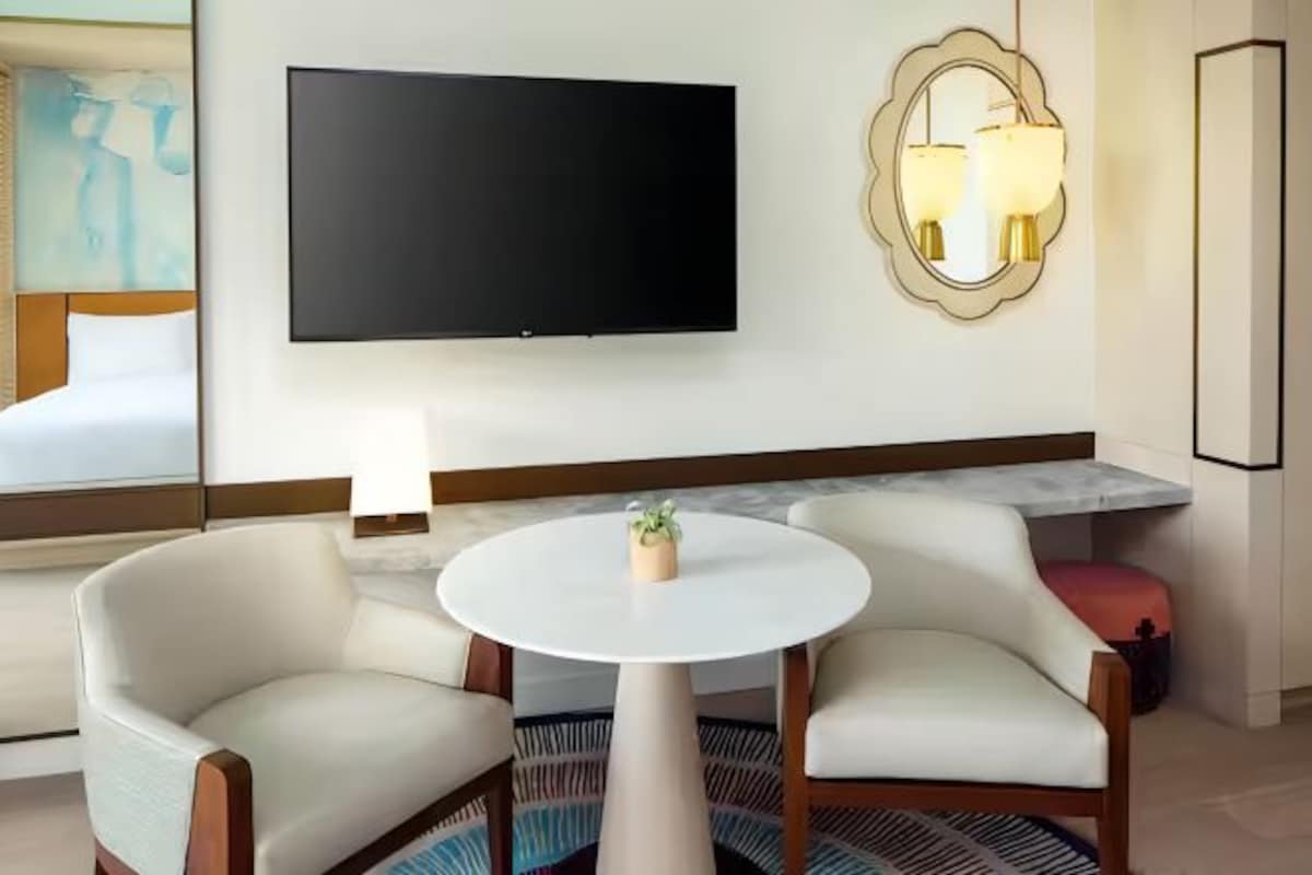 A hotel room with a large flat-screen TV mounted on the wall, two white armchairs around a small round table, a bedside table with a lamp, and a large mirror above the mirror.