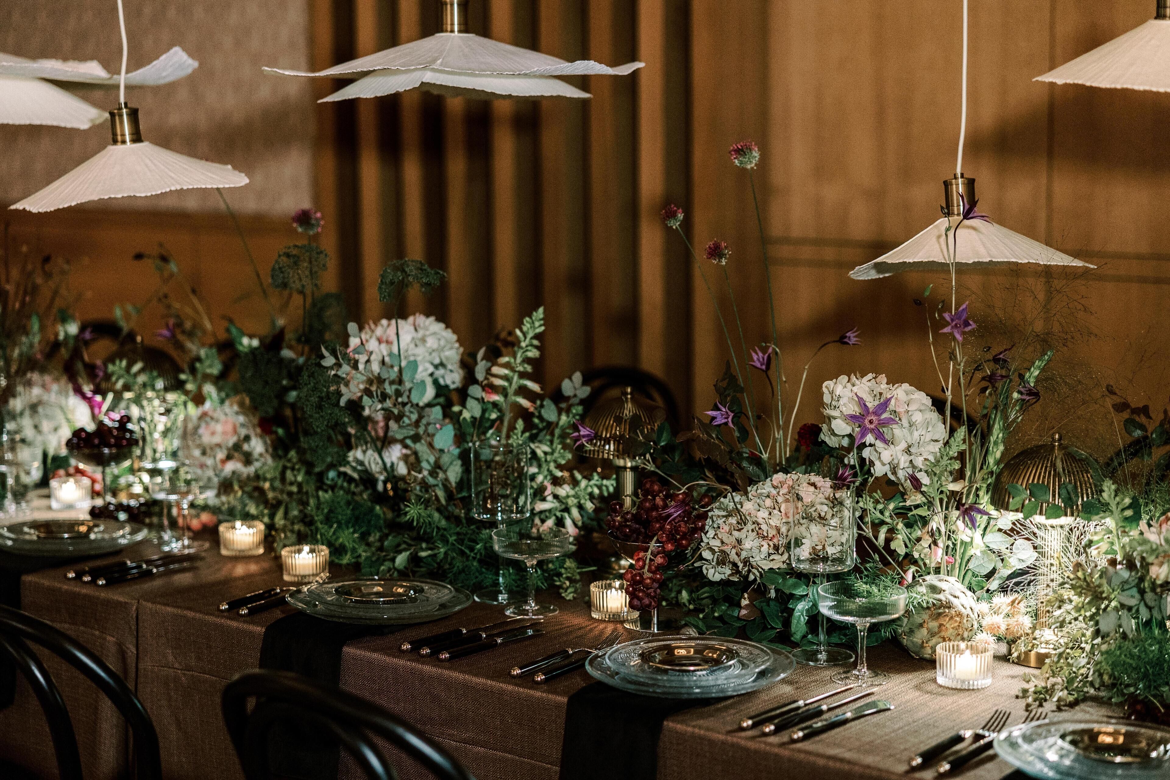 Een eettafel gedekt met borden, bestek en kaarsen, omringd door bloemdecoraties en hanglampen.