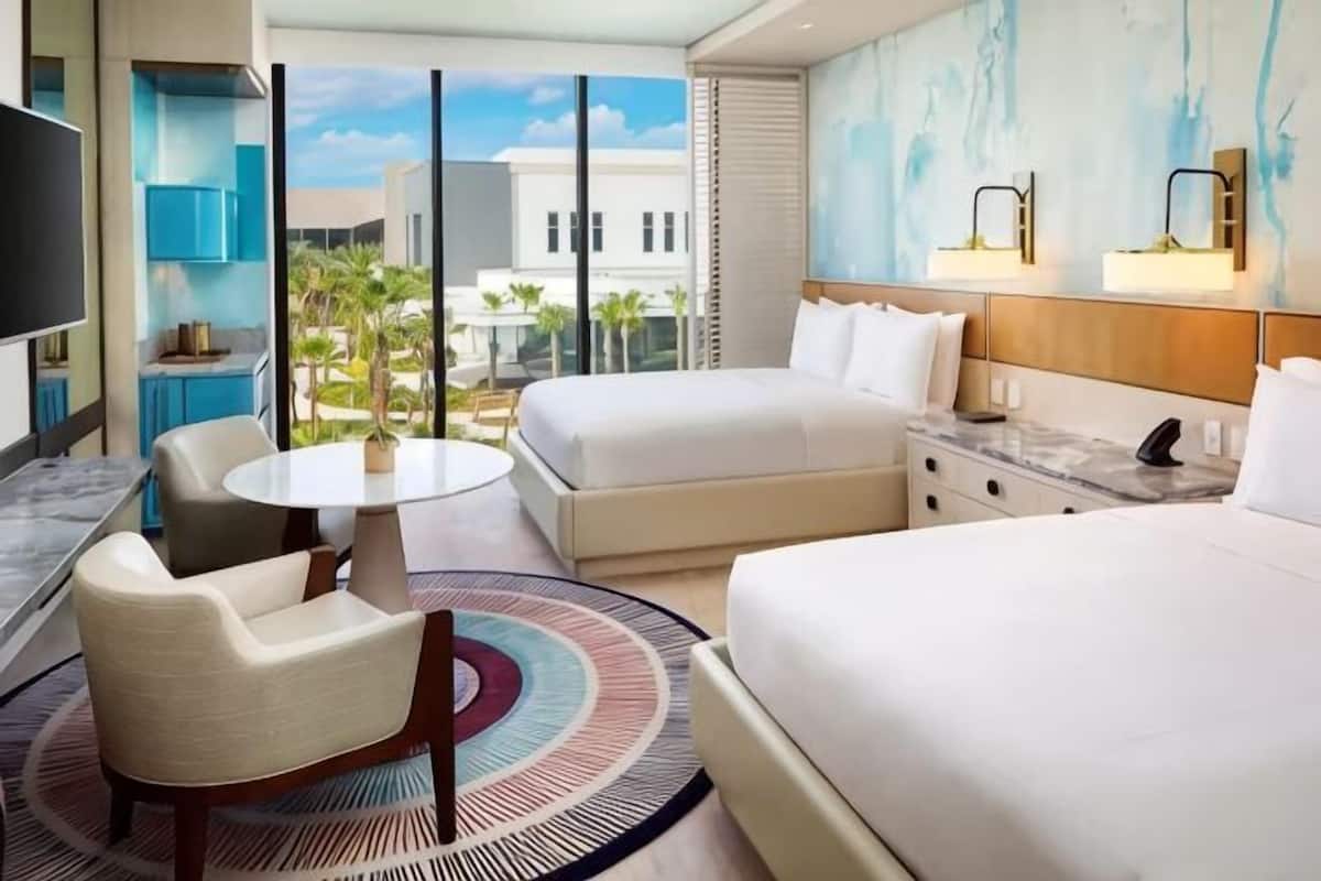 Une chambre d’hôtel avec un lit, un bureau, une chaise, une petite table et une grande fenêtre donnant sur des immeubles et des palmiers.