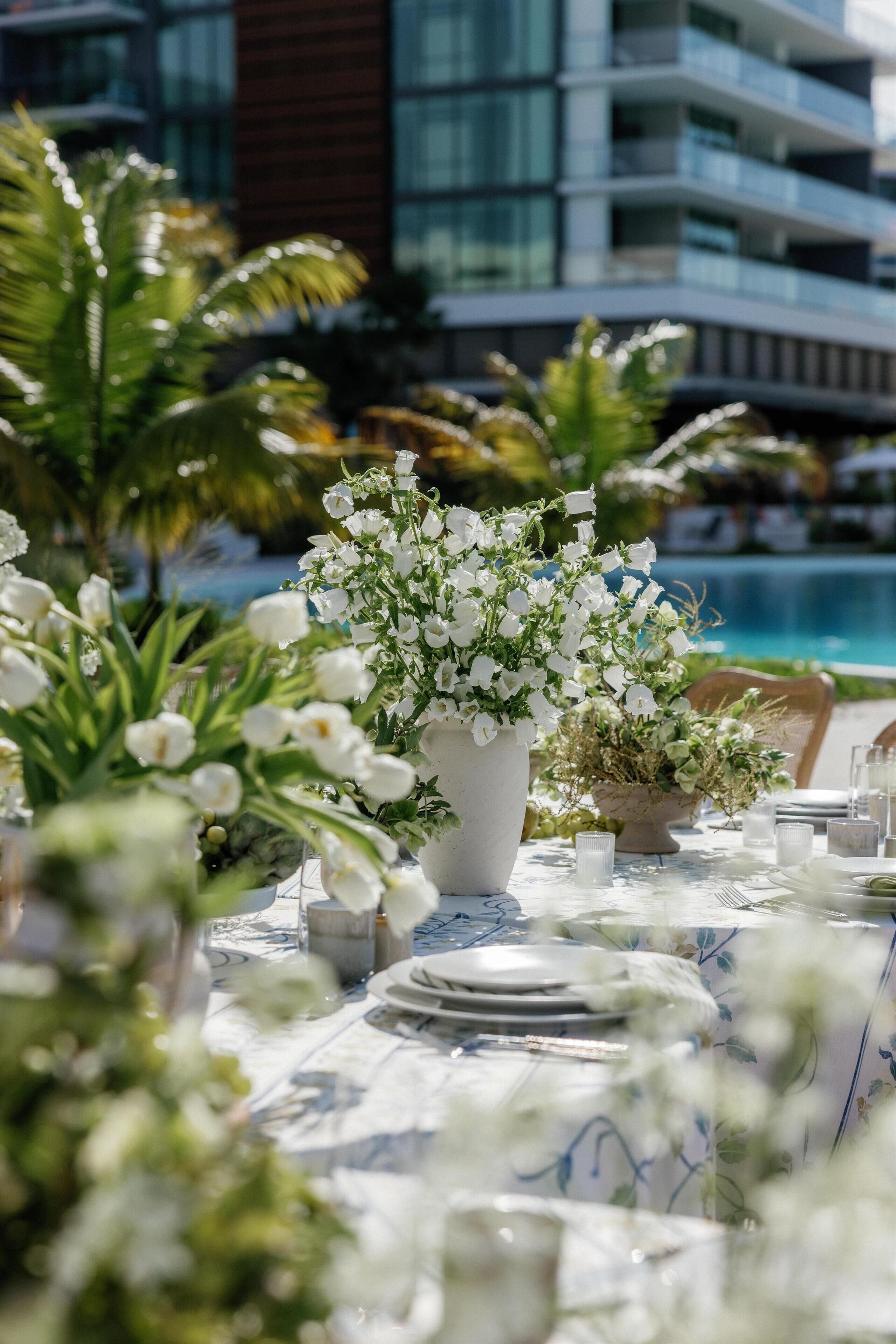 A dining table set for a meal with white flowers and a view of a building and pool in the background.