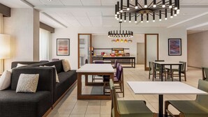 A modern hotel lobby with a sofa, dining tables, and a kitchen area in the background.