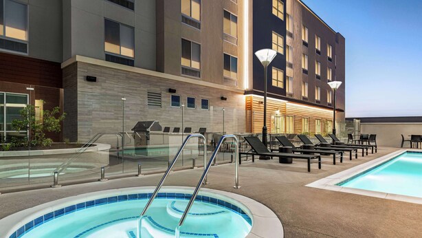 A rooftop pool area with lounge chairs and a hot tub, surrounded by a modern building with large windows.