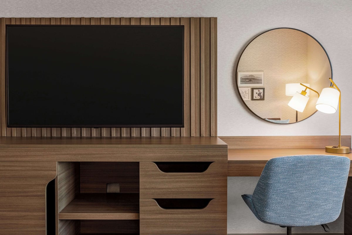 Moderni hotellihuone, jossa on puinen työpöytä, sininen tuoli, suuri litteänäyttöinen televisio ja pyöreä peili, jossa on valaisin.