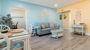 A living room with a light blue wall, a grey sofa, a glass coffee table, and a white dining table with chairs.