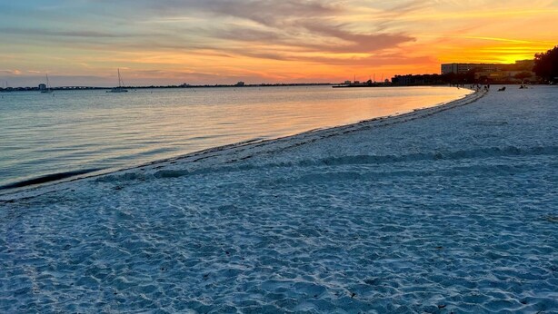 A beach with a sunset, calm water, and a distant shoreline with buildings.