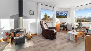 Een gezellige woonkamer met een open haard, een leren fauteuil, een houten tafel en een groot raam met uitzicht op de bergen.