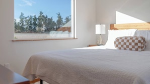 Een slaapkamer met een groot raam, een bed met een geruit kussen, een nachtkastje met een lamp en een houten nachtkastje.