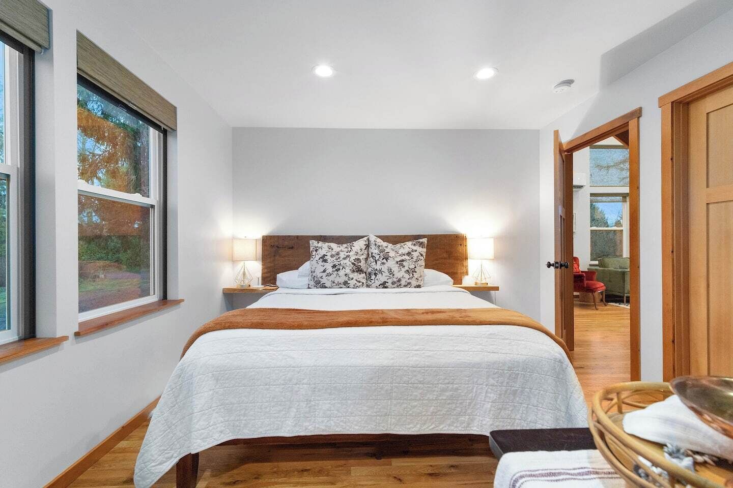 A bedroom with a large bed, two bedside tables with lamps, a wooden headboard, and a window with a view of trees.