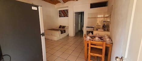 A room with a bed, a small table with cups, a kitchen area, and a door.
