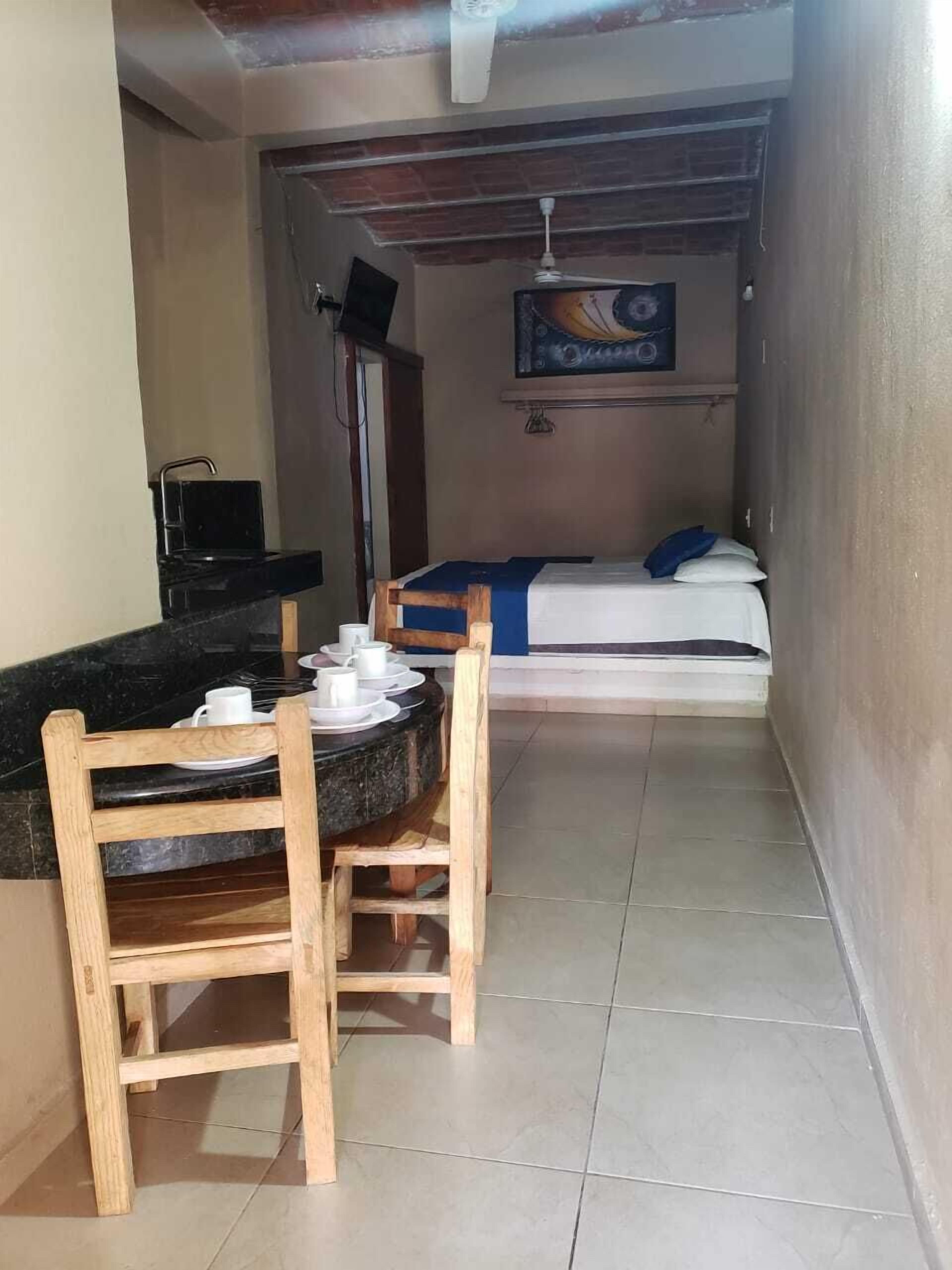 A compact room with a bed, a small table with chairs, a painting on the wall, and a ceiling fan.