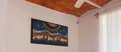Una cama individual con colcha y almohadas azules, un ventilador de techo, un cuadro en la pared y una ventana con cortinas.