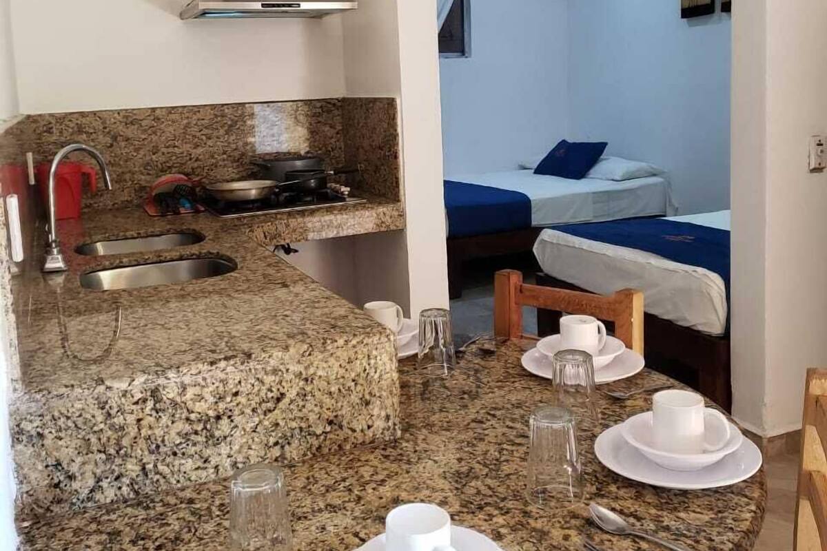 A compact kitchen with granite countertops, a stove, and a sink. There is a dining area with a table set for two, and a bed with a blue bedspread in the background.