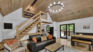 A spacious living area with a wooden staircase, a black leather sofa, a wooden dining table, and a large window offering a view of the outdoors.