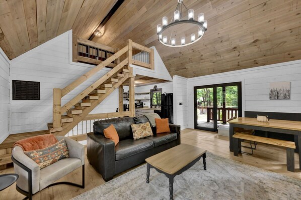 A spacious living area with a wooden staircase, a black leather sofa, a wooden dining table, and a large window offering a view of the outdoors.