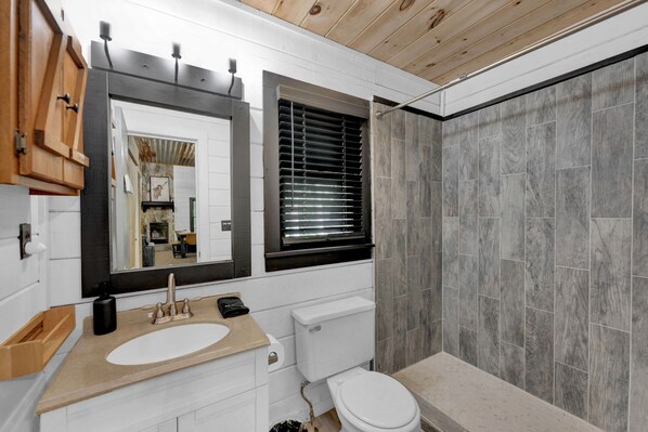 A modern bathroom with a wooden ceiling, a large mirror, a sink with a beige countertop, a toilet, and a shower area with grey tiles.