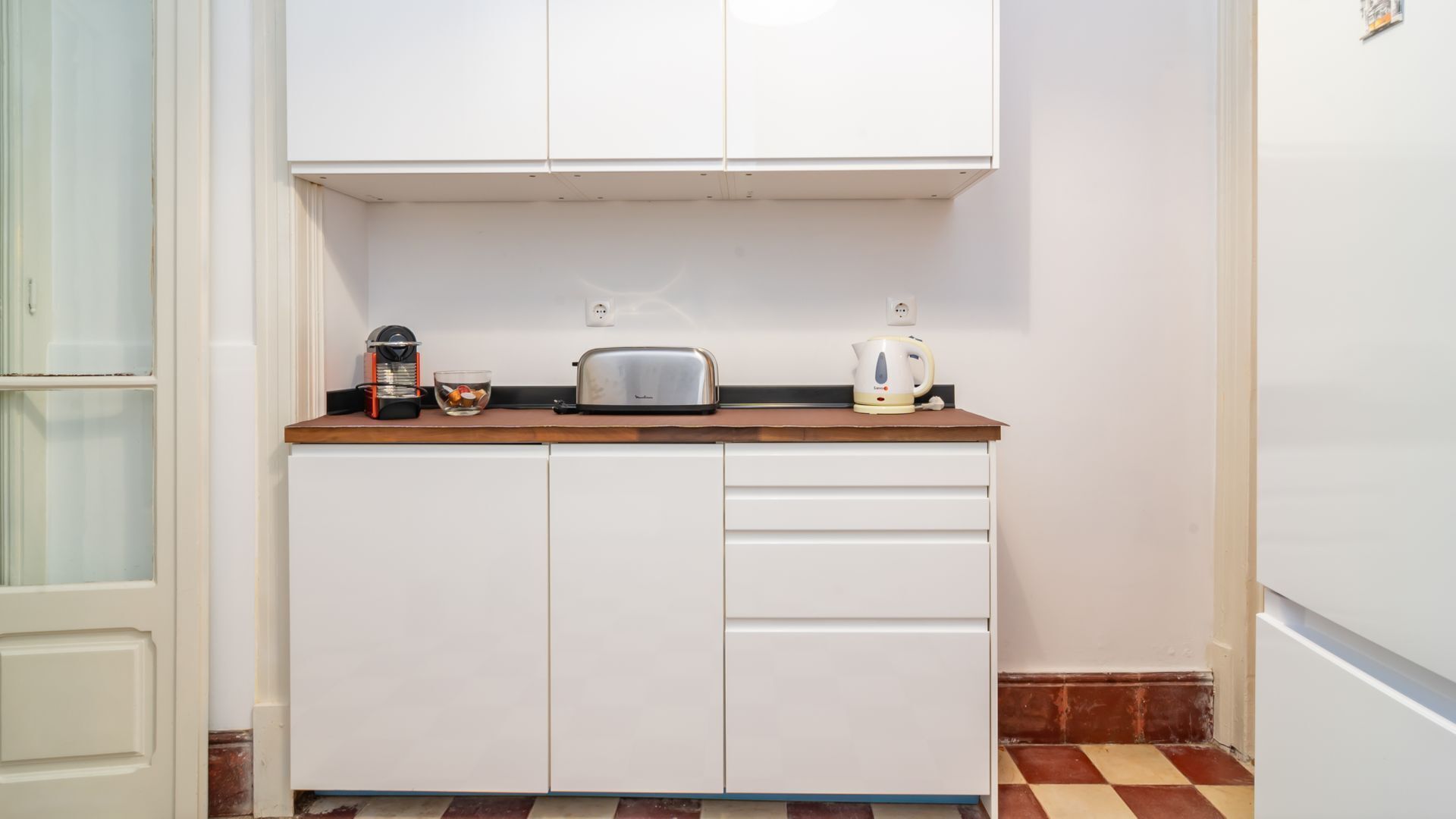 A compact kitchen with white cabinets, a toaster, a kettle, and a coffee maker on the countertop.