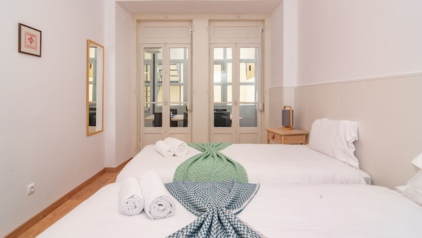 Habitación de hotel con dos camas, piso de madera, una mesita de noche pequeña con lámpara y vista a través de un ventanal.