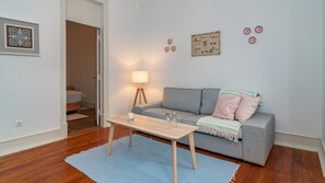 Un salon moderne avec un canapé gris, une table basse en bois et un tapis bleu. On y trouve une lampe, un tableau encadré et une petite table ornée d’objets décoratifs.