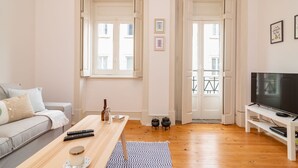 Ein Wohnzimmer mit einer Couch, einem Couchtisch, einem Fernseher auf einem Schrank und einem Balkon mit Geländer.