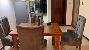 A dining area with a wooden table and four upholstered chairs. A refrigerator and kitchen area are visible in the background. - New Luxury Apartment very secure at State House Nairobi (kilimani)