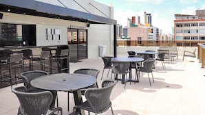 An outdoor patio area with multiple tables and chairs, a bar area with a sign, and a cityscape in the background. - Apart no Mardisa Design por Carpediem (João Pessoa)