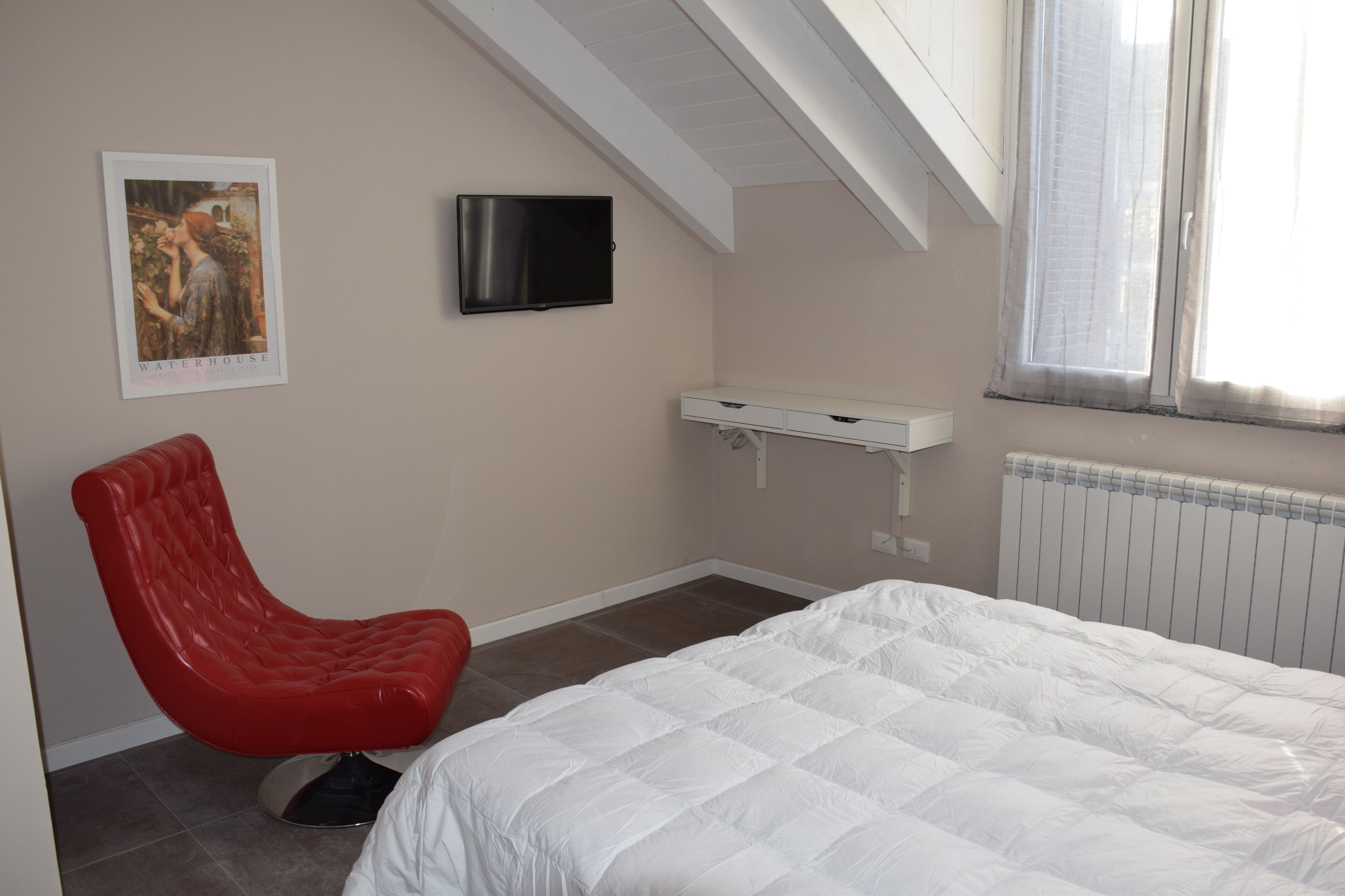 A bedroom with a bed, a red chair, a TV, a small white desk, and a radiator.