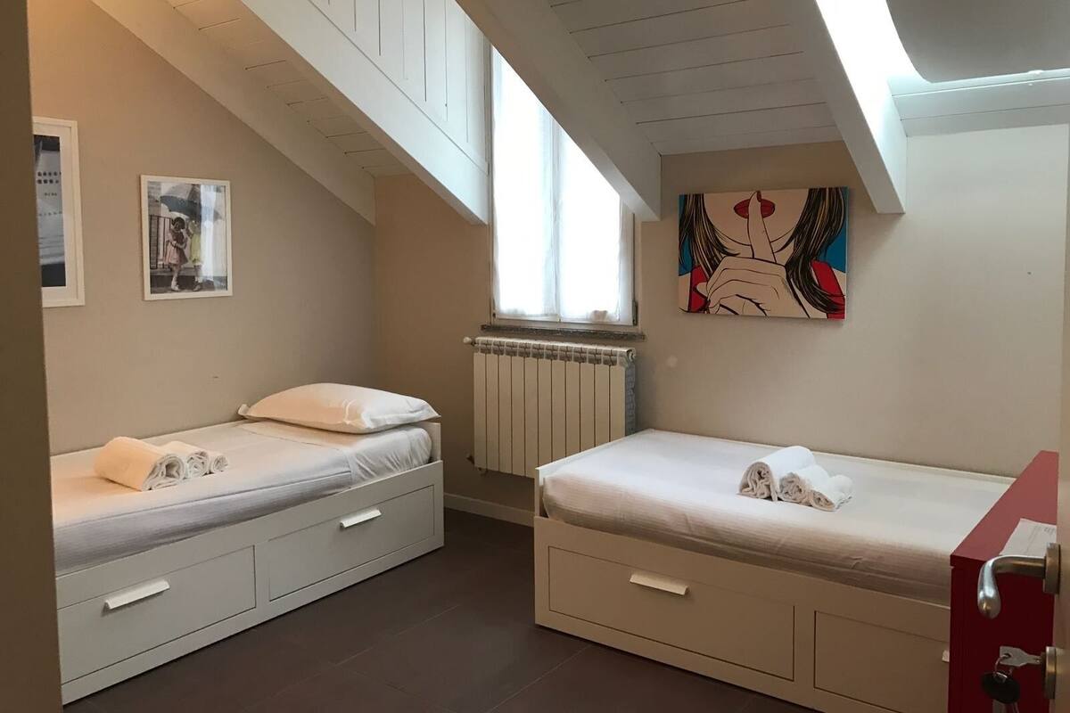 A small, well-lit room with two single beds, a radiator, a window, and a painting on the wall.