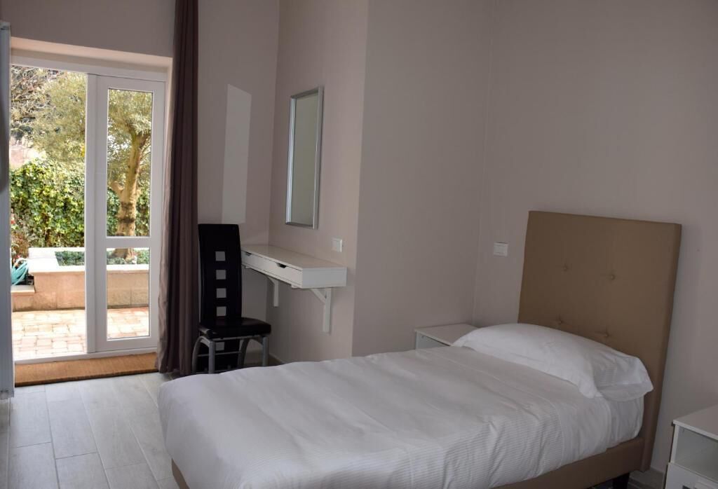 A bedroom with a bed, a desk, a chair, a mirror, and a sliding glass door to a balcony.
