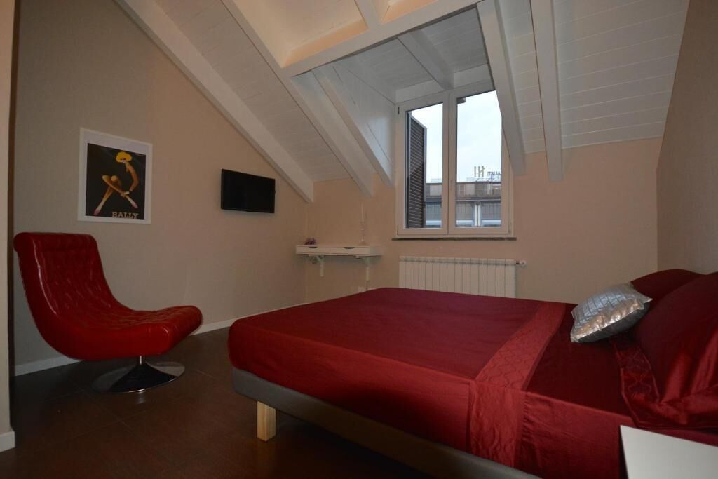A bedroom with a red bed, a red chair, a small window, and a wall-mounted TV.