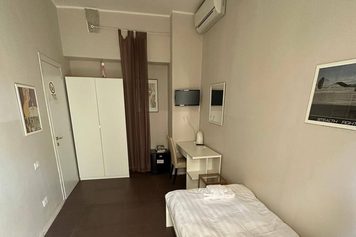 A hotel room with a bed, a desk, a chair, a wardrobe, a TV, and an air conditioning unit.