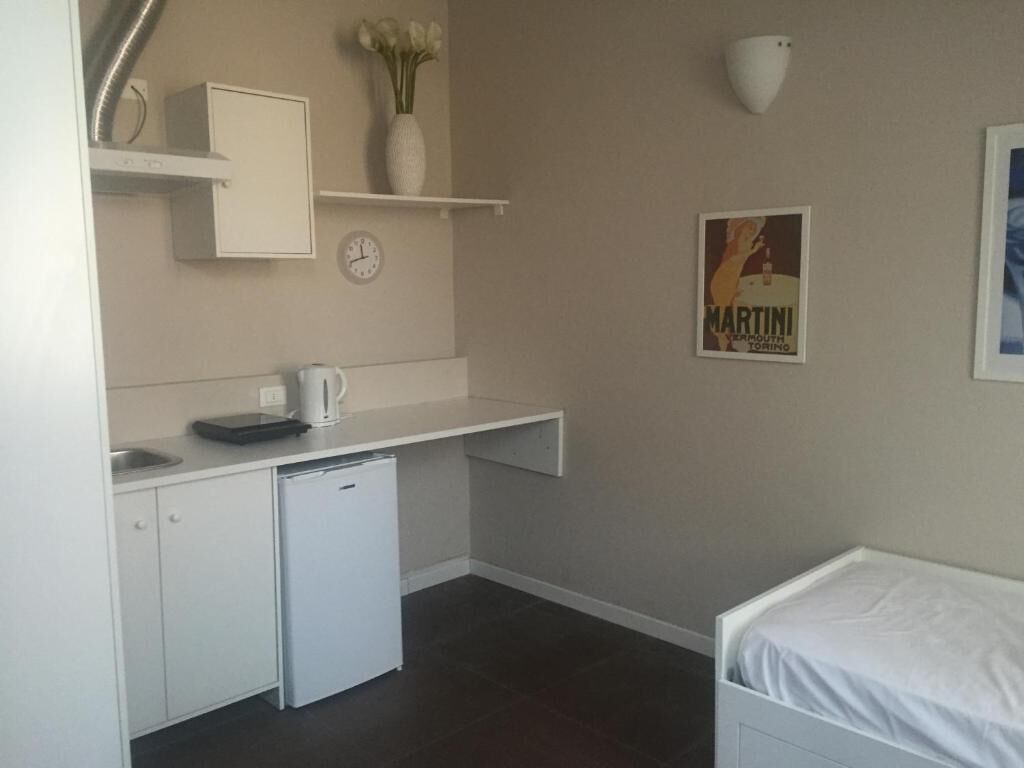 Ein kleines Zimmer mit einer Küchenzeile, einem Bett, einem kleinen weißen Schrank, einer Uhr und einem Poster an der Wand.