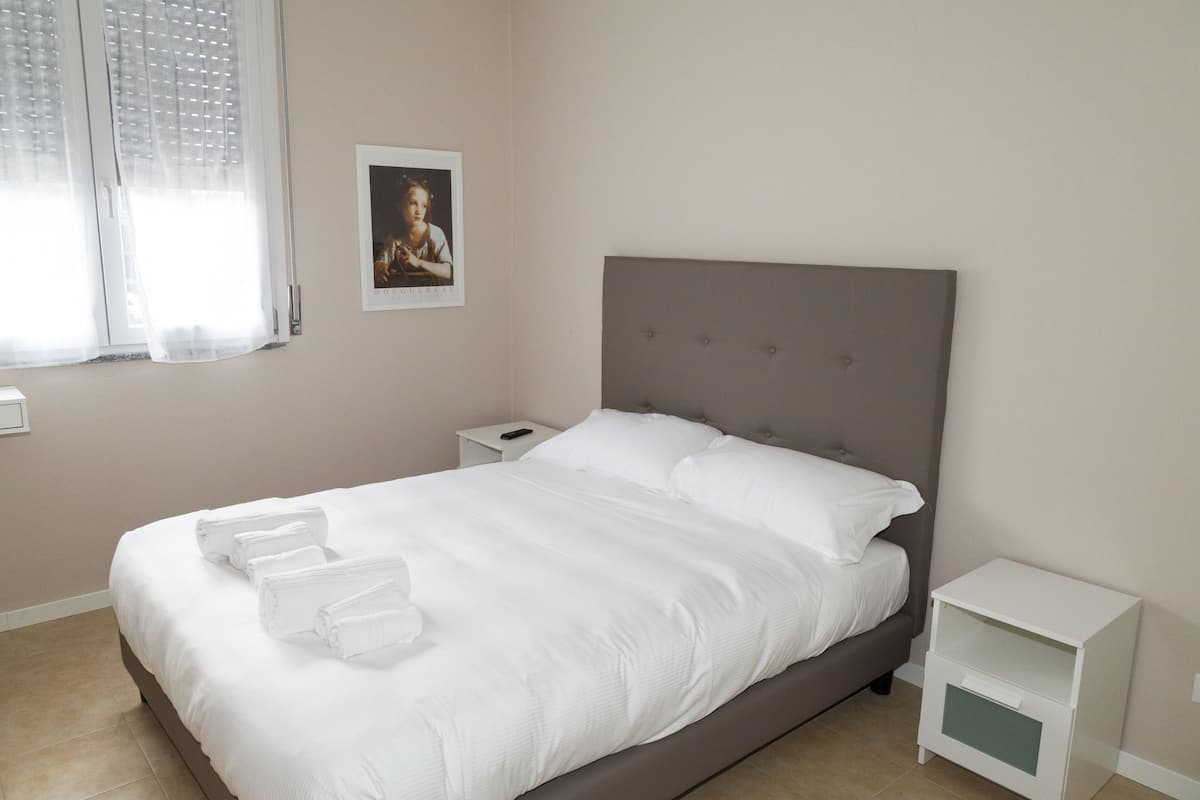 A neatly made bed with white linens and towels, a bedside table with a lamp, and a framed picture on the wall.