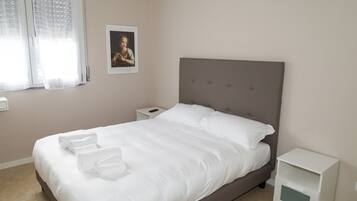 Ein ordentlich bezogenes Bett mit weißen Bettwäsche und Handtüchern, ein Nachttisch mit Lampe sowie ein gerahmtes Bild an der Wand.
