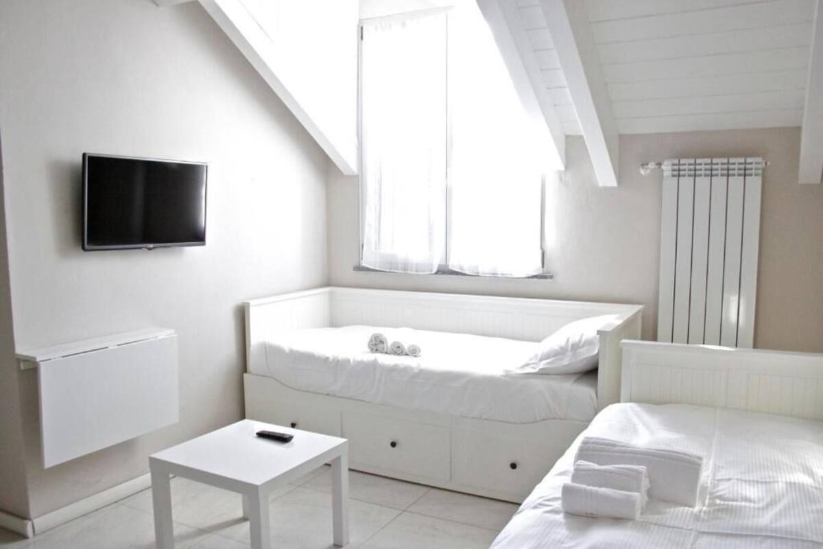 A modern, white-themed bedroom with a flat-screen TV, a small bedside table, a window with curtains, and a radiator.