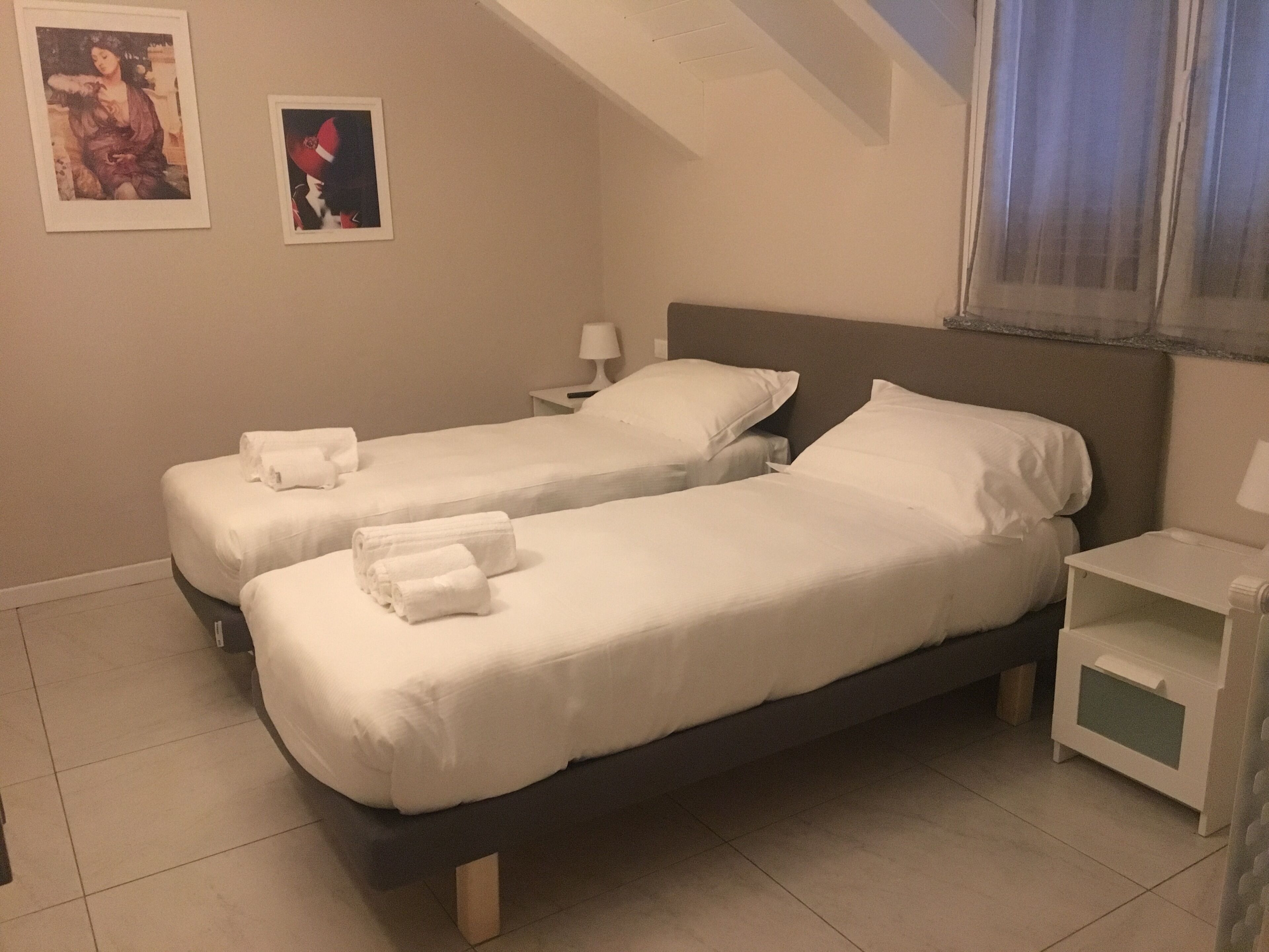 A hotel room with two single beds, each with white linens and towels, a nightstand with a lamp, and a small shelf.