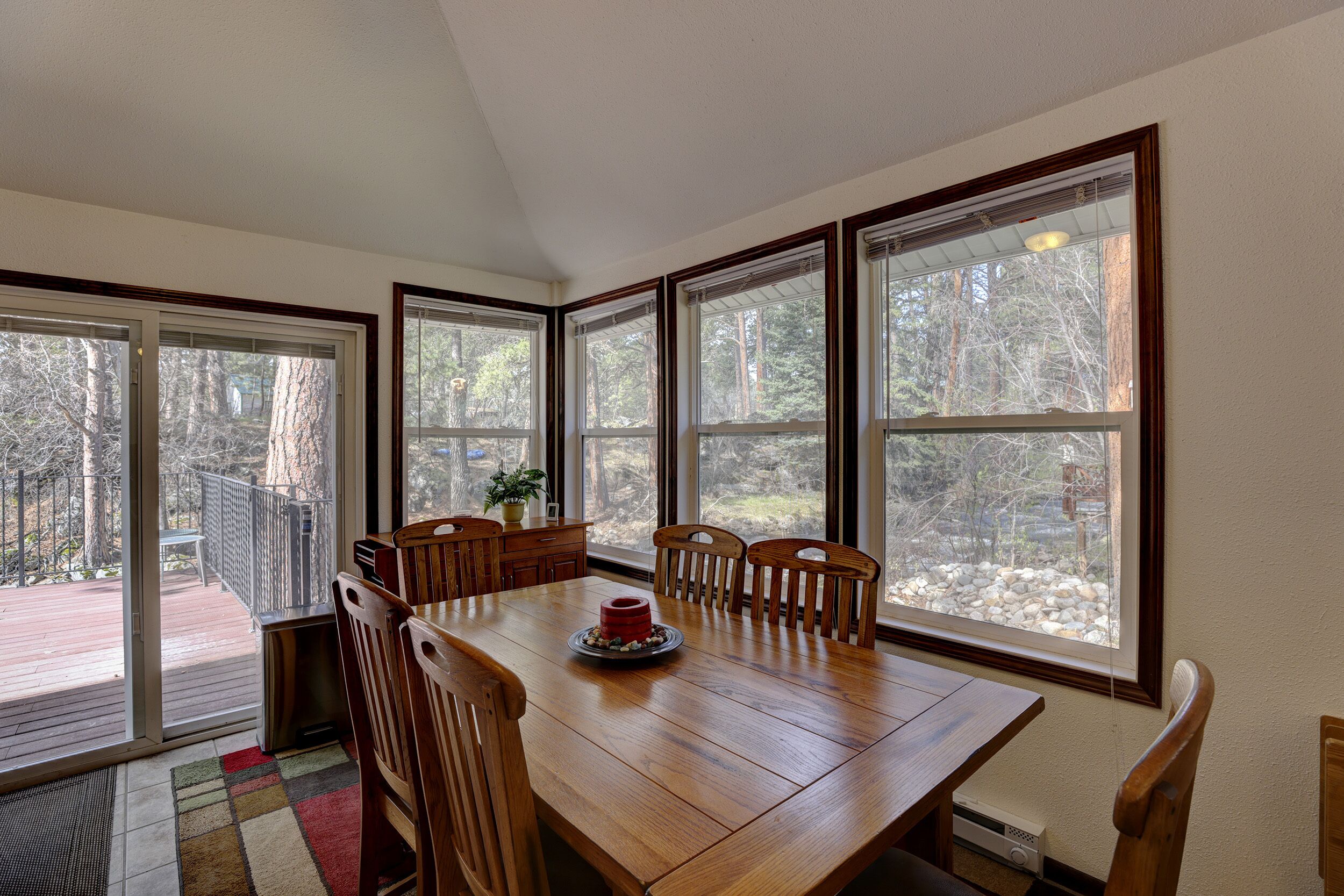 A dining area with a wooden table and chairs, large windows, and a view of a deck and trees.